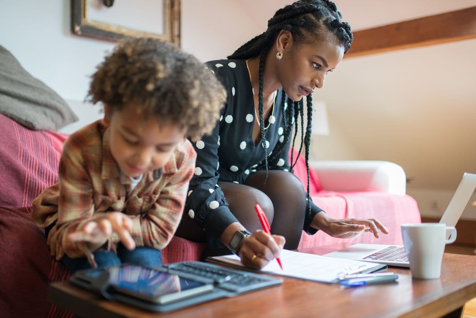 A mother fills out a school form at home while her young son leans against her